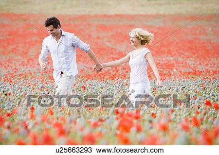 Stock Image - Couple walking in poppy field holding hands smiling. Fotosearch