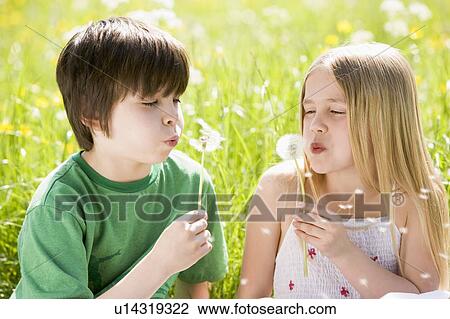 Stock Image - Two young children sitting outdoors blowing dandelion heads smiling. Fotosearch