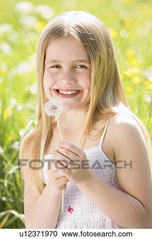 Stock Image - Young girl sitting outdoors holding dandelion head smiling. Fotosearch