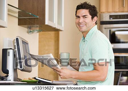 Stock Photo - Man in kitchen with computer holding newspaper and coffee smiling. Fotosearch