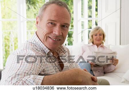 Stock Photograph - Man in living room smiling with woman in background reading newspaper. Fotosearch