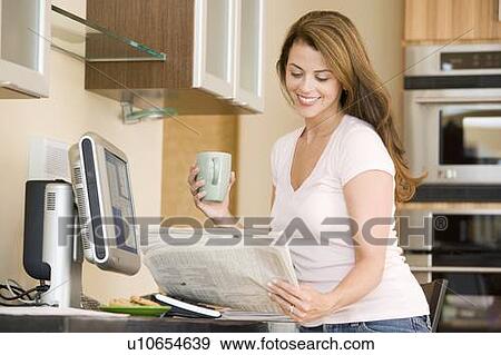 Stock Photo - Woman in kitchen at computer with newspaper and coffee smiling. Fotosearch
