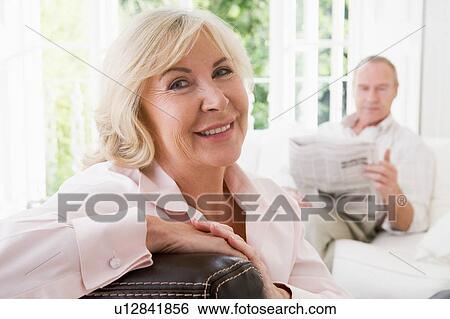 Stock Photograph - Woman in living room smiling with man in background reading newspaper. Fotosearch