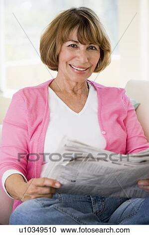 Stock Image - Woman relaxing with newspaper in living room and smiling. Fotosearch
