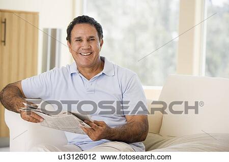 Stock Image - Man in living room reading newspaper smiling. Fotosearch