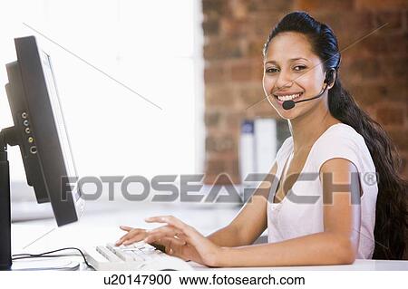 Stock Image - Businesswoman in office wearing headset and typing on computer smiling. Fotosearch