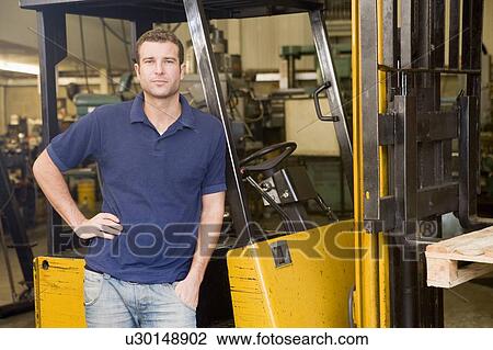 Stock Image - Warehouse worker standing by forklift. Fotosearch