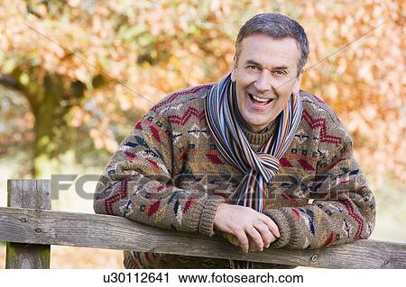 Stock Image - Man outdoors at park leaning on fence smiling (selective focus). Fotosearch