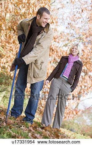 Picture - Man outdoors raking leaves and woman in background smiling (selective focus). Fotosearch