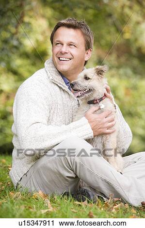 Stock Image - Man sitting outdoors with dog in park smiling (selective focus). Fotosearch