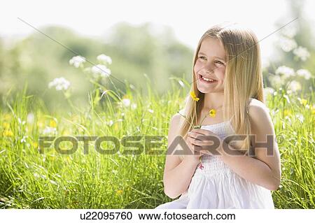 Stock Image - Young girl sitting outdoors holding flower smiling. Fotosearch