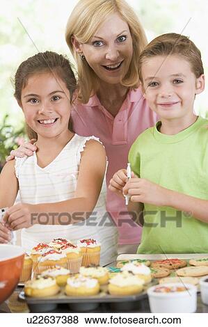 Stock Photo - Woman with two children in kitchen decorating cookies smiling. Fotosearch