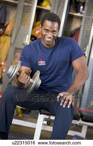 Stock Photograph - Fireman lifting dumbbell on bench in fire station locker room. Fotosearch