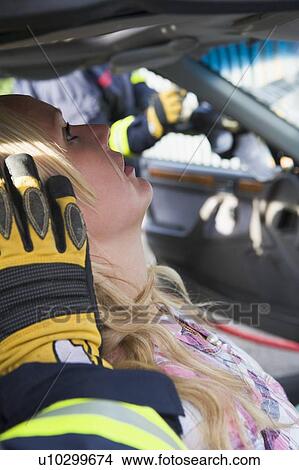 Picture - Injured woman in car with firefighter in background cutting out windshield (selective focus). Fotosearch