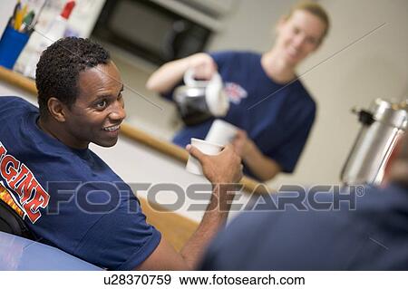 Stock Photo - Three firefighters in break room with one getting coffee in background (selective focus). Fotosearch