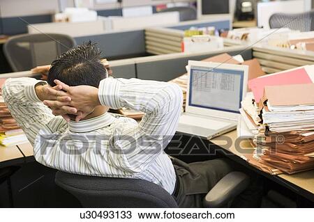 Stock Image - Businessman in cubicle with laptop and stacks of files. Fotosearch