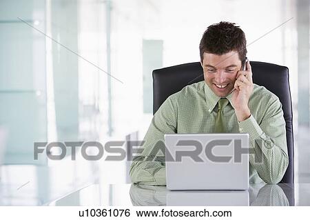 Businessman sitting in office using cellular phone and laptop View Large Photo Image Stock Photograph - Businessman sitting in office using cellular phone and laptop. Fotosearch