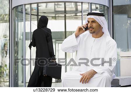 Businessman sitting outdoors by building using cellular phone with businesswoman walking in background (selective focus) View Large Photo Image Stock Photo - Businessman sitting outdoors by building using cellular phone with businesswoman walking in background (selective focus). Fotosearch