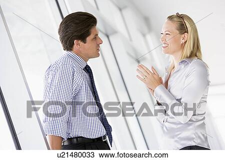 Stock Image - Businessman standing in corridor talking to smiling businesswoman. Fotosearch