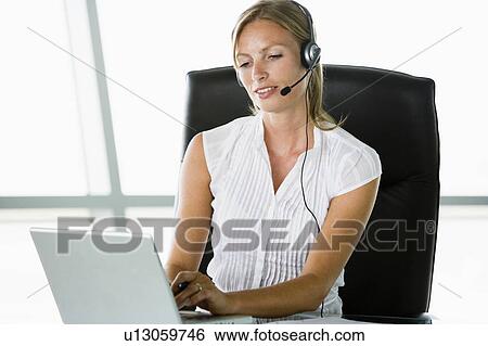 Businesswoman sitting in office wearing headset using laptop View Large Photo Image Stock Photograph - Businesswoman sitting in office wearing headset using laptop. Fotosearch