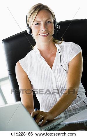 Stock Photograph - Businesswoman sitting in office wearing headset using laptop and smiling. Fotosearch