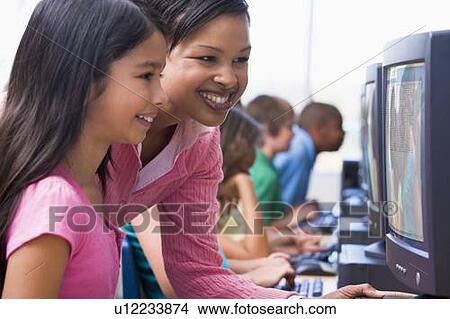 Picture - Teacher helping student at computer terminal with students in background (depth of field/high key). Fotosearch