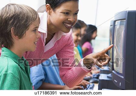 Stock Image - Teacher helping student at computer terminal with students in background (depth of field/high key). Fotosearch