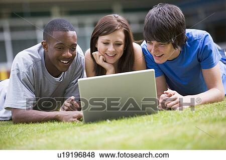 Stock Photo - Three students lying outdoors on lawn with laptop. Fotosearch