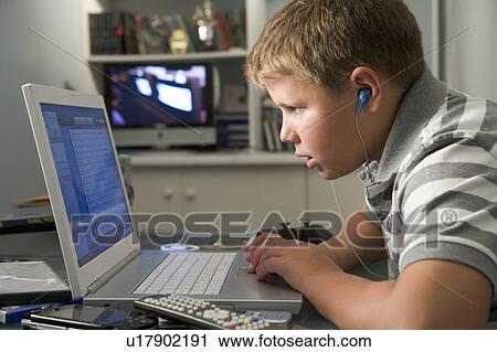 Stock Image - Young boy in bedroom using laptop and listening to MP3 player. Fotosearch