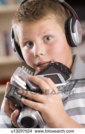 Picture - Young boy wearing headphones in bedroom holding many electronic devices. Fotosearch