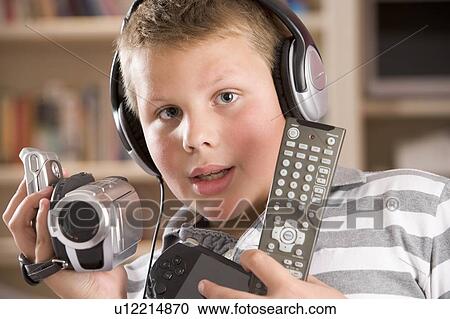 Stock Image - Young boy wearing headphones in bedroom holding many electronic devices. Fotosearch