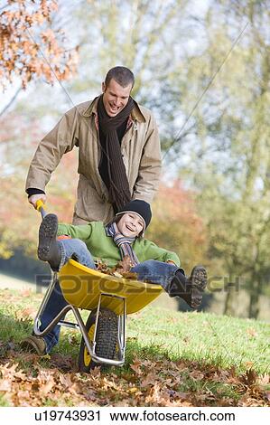 Stock Image - Man and young boy outdoors playing with wheelbarrow and smiling (selective focus). Fotosearch