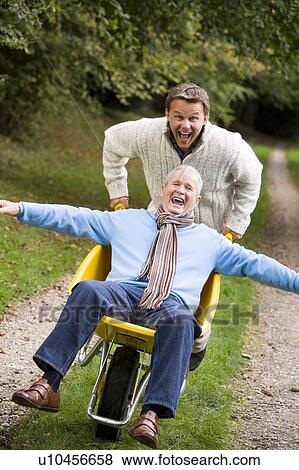 Man walking on path outdoors pushing other man in wheelbarrow and smiling (selective focus) View Large Photo Image Stock Photo - Man walking on path outdoors pushing other man in wheelbarrow and smiling (selective focus). Fotosearch