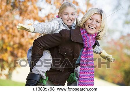 Stock Photo - Mother outdoors piggybacking daughter and smiling (selective focus). Fotosearch