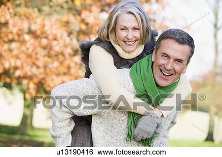 Stock Photograph - Senior man outdoors piggybacking woman and smiling (selective focus) . Fotosearch