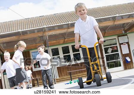 Stock Image - Students playing outdoors during recess one with a tricycle scooter (selective focus). Fotosearch