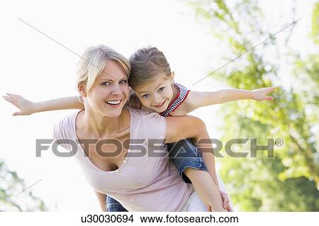 Picture - Woman giving young girl piggyback ride outdoors smiling. Fotosearch