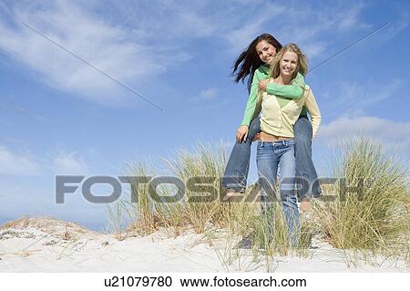 Young woman giving piggyback ride to her friend on a sand hill View Large Photo Image Stock Image - Young woman giving piggyback ride to her friend on a sand hill. Fotosearch