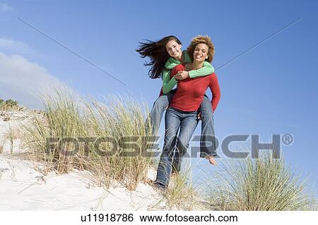 Young woman giving piggyback ride to her friend on a sand hill View Large Photo Image Stock Photograph - Young woman giving piggyback ride to her friend on a sand hill. Fotosearch