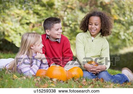 Stock Photo - Three young friends sitting on grass with pumpkins smiling. Fotosearch