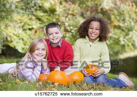 Stock Photography - Three young friends sitting on grass with pumpkins smiling. Fotosearch