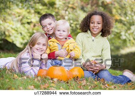 Picture - Three young friends with baby sitting on grass with pumpkins smiling. Fotosearch