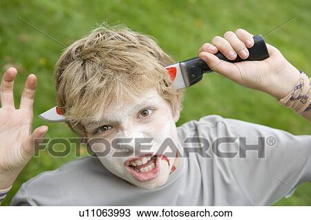 Stock Image - Young boy with scary Halloween make up and plastic knife through head. Fotosearch