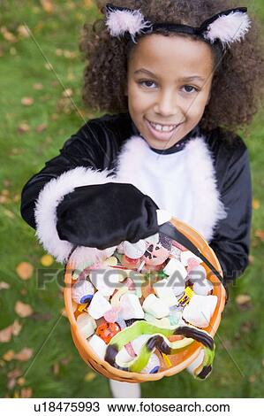 Stock Image - Young girl outdoors in cat costume on Halloween holding candy. Fotosearch