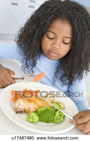 Stock Image - Young girl in kitchen eating chicken and vegetables. Fotosearch