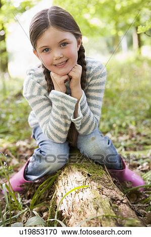 Stock Image - Young girl outdoors in woods sitting on log smiling. Fotosearch