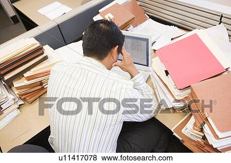 Stock Photo - Businessman in cubicle with laptop and stacks of files. Fotosearch