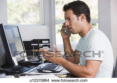 Picture - Man in home office on telephone using computer and frowning. Fotosearch