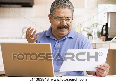 Stock Image - Man in kitchen with laptop and paperwork frustrated. Fotosearch
