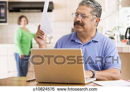 Stock Photograph - Man in kitchen with laptop and paperwork with woman in background. Fotosearch